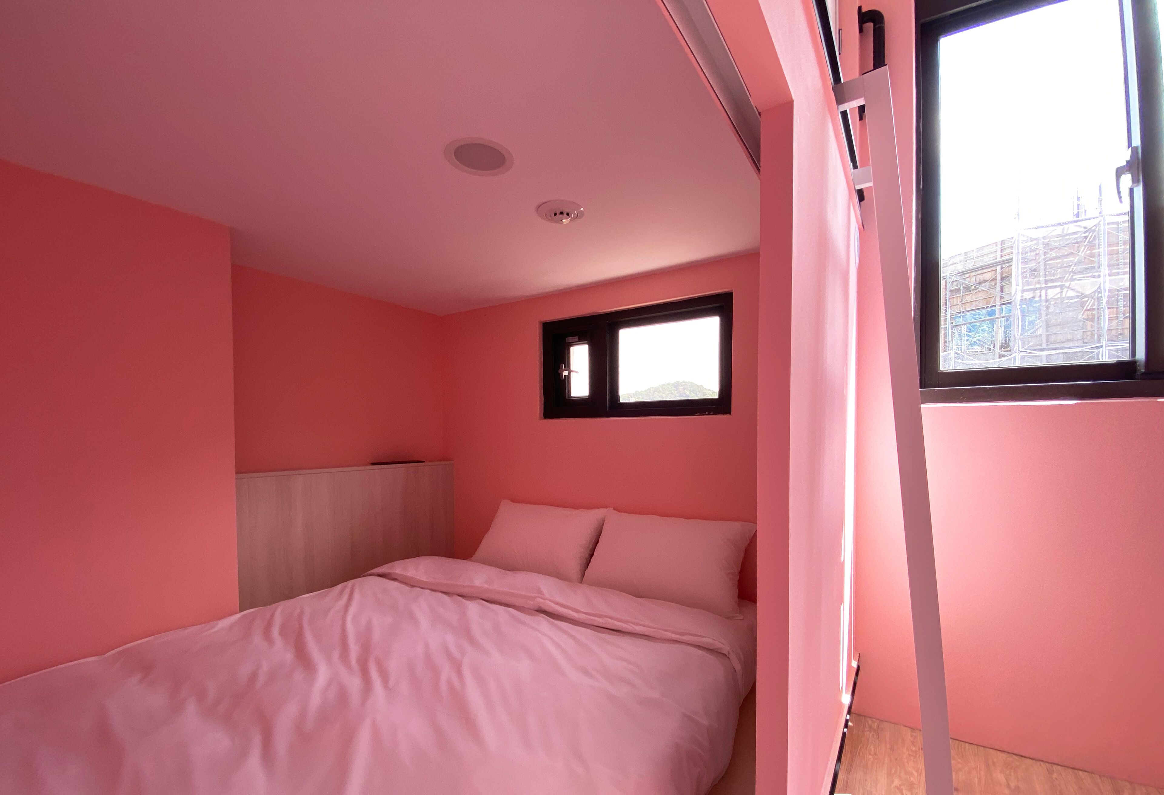 Un dormitorio decorado en tonos rosados, con cama, ventana y vista a un edificio.