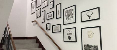 Un escalier avec une main courante en bois mène à un palier où plusieurs œuvres d’art encadrées sont exposées sur le mur.