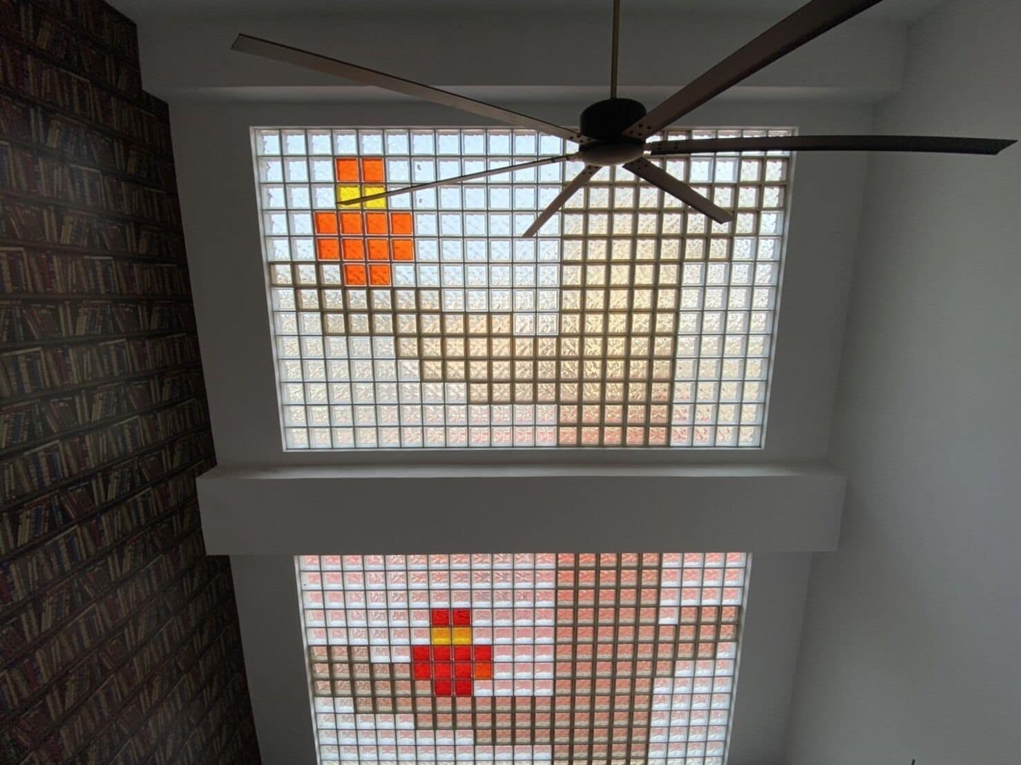 A room with a ceiling fan, a bookshelf, and two windows with grid patterns.