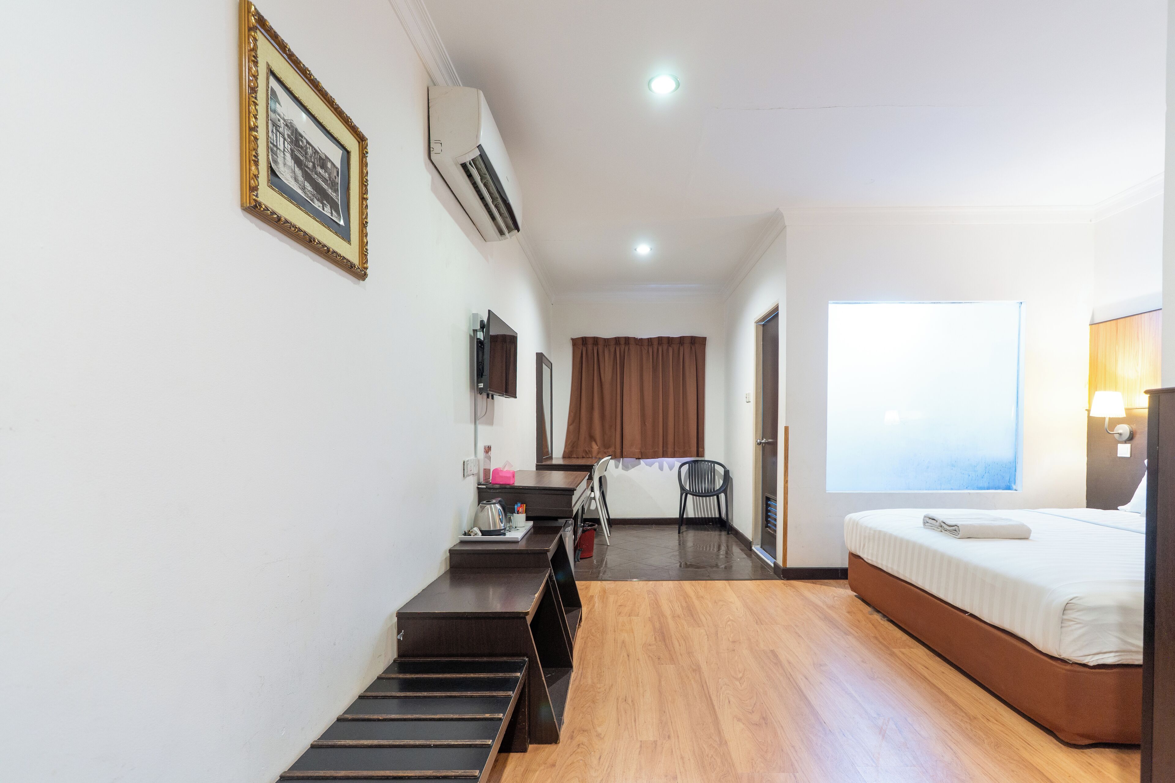 A hotel room with a bed, a desk with a chair, an air conditioner, a window with curtains, and a picture frame on the wall.