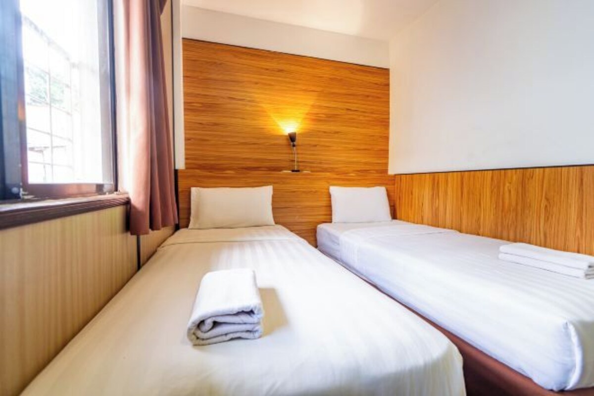 A hotel room with two beds, wooden headboard, a wall-mounted light, and a window with curtains.