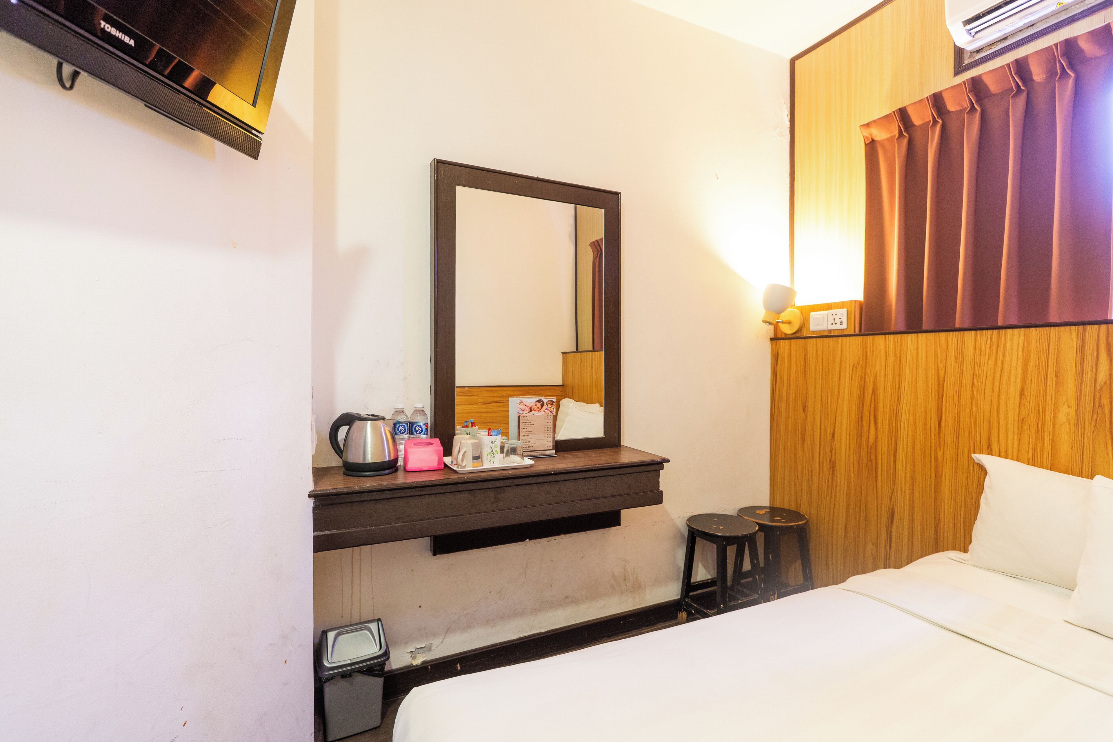 Ein Hotelzimmer mit einem Bett, einem Holz-Kopfteil, einem Spiegel, einem kleinen Tisch mit Wasserkocher, Toaster und einer Wasserflasche.