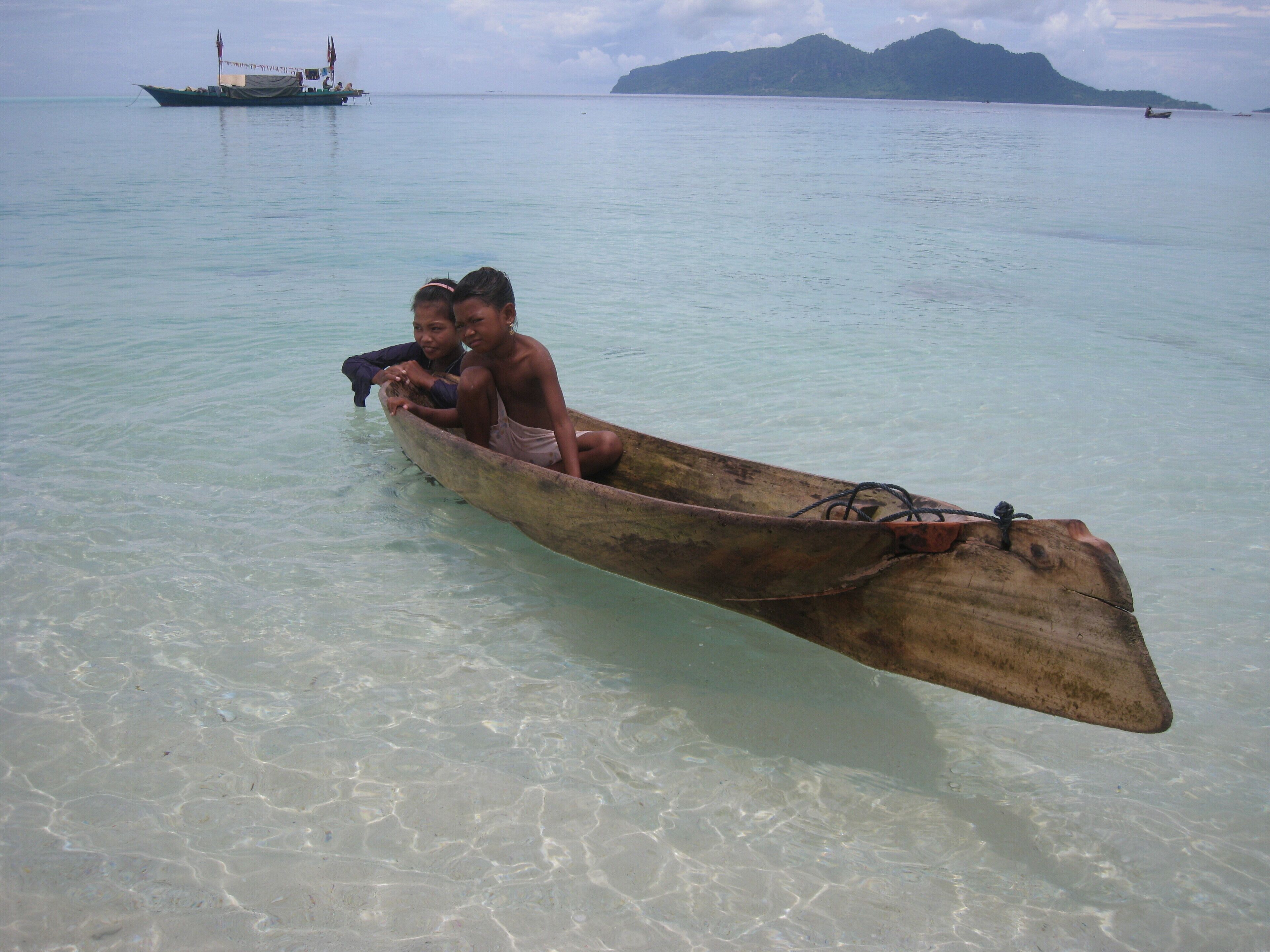 Un bateau en bois avec deux personnes à bord, naviguant dans des eaux peu profondes, un autre bateau plus éloigné, et une montagne en arrière-plan.