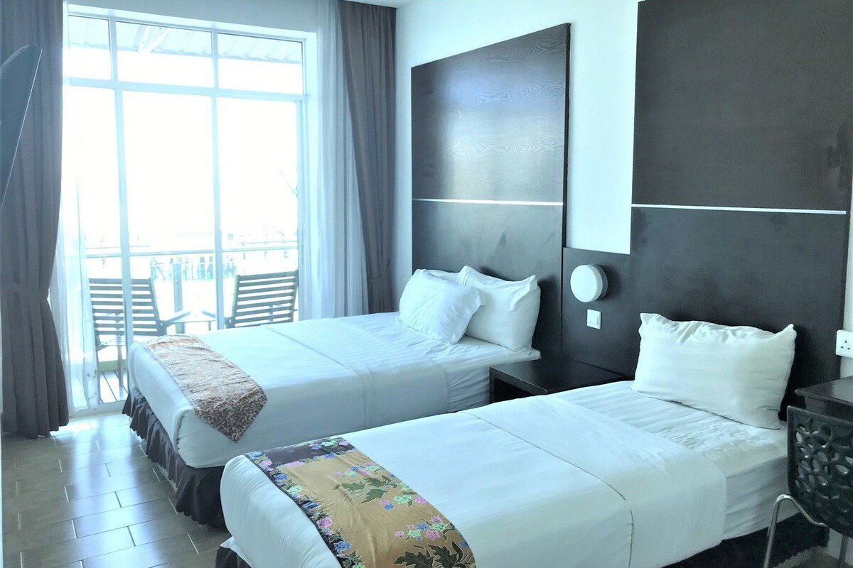 Habitación de hotel con dos camas, un ventanal con cortinas y vistas al exterior, además de una pequeña mesa con una silla.