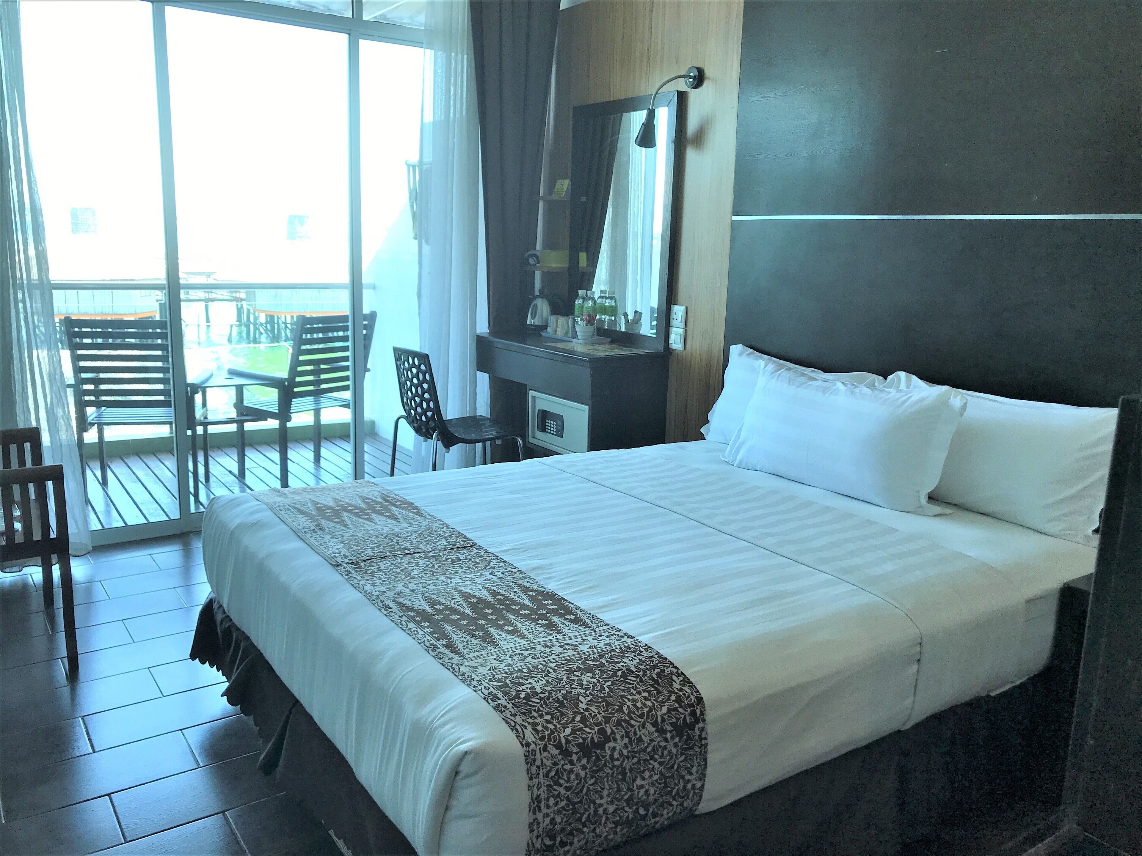 A hotel room with a large bed, a desk with a mirror, and a chair. There is a balcony with chairs and a table.
