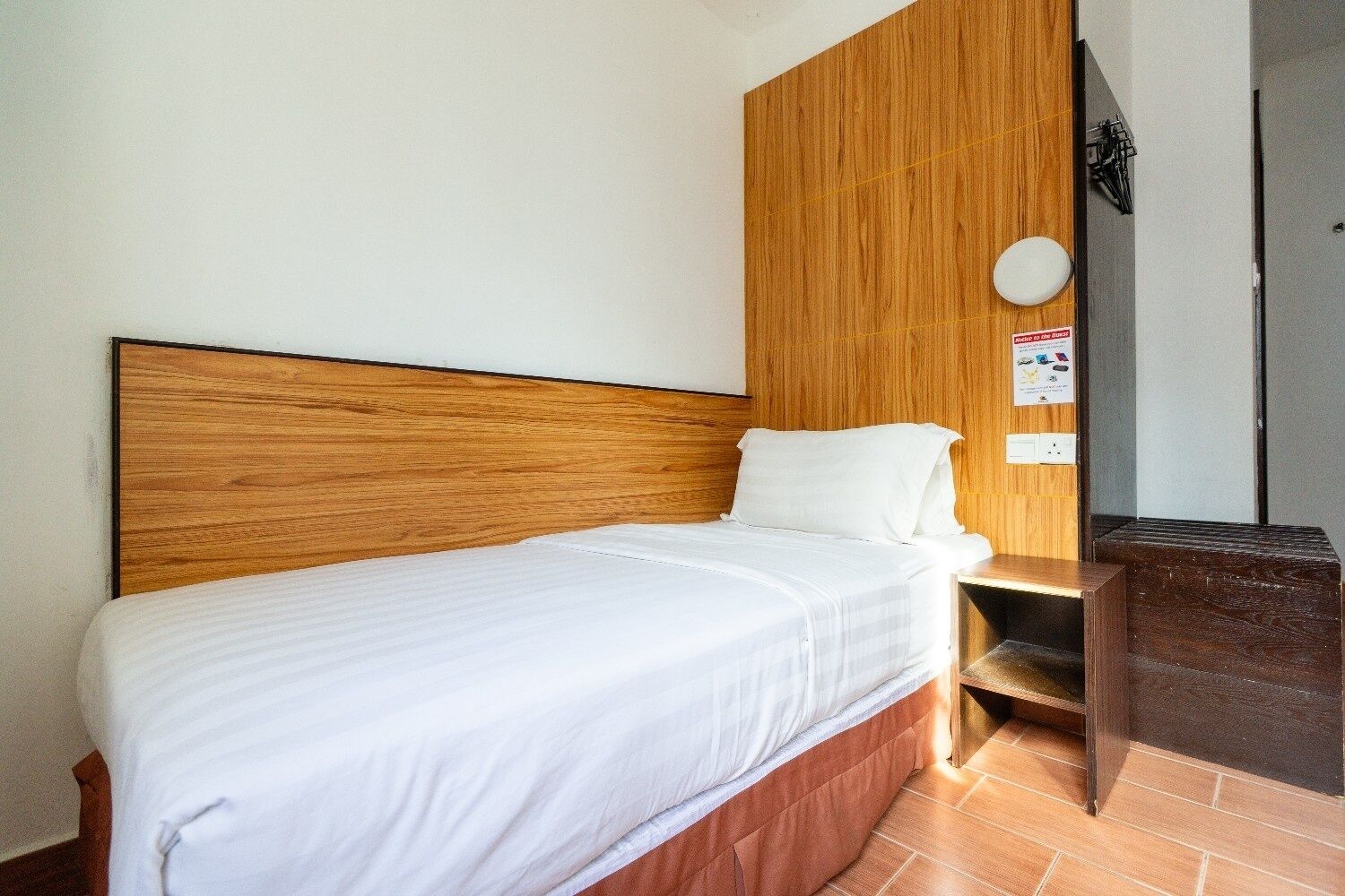 Una habitación de hotel con cabecera de madera, una cama individual con ropa blanca, una mesita de noche de madera y un cubo de basura.