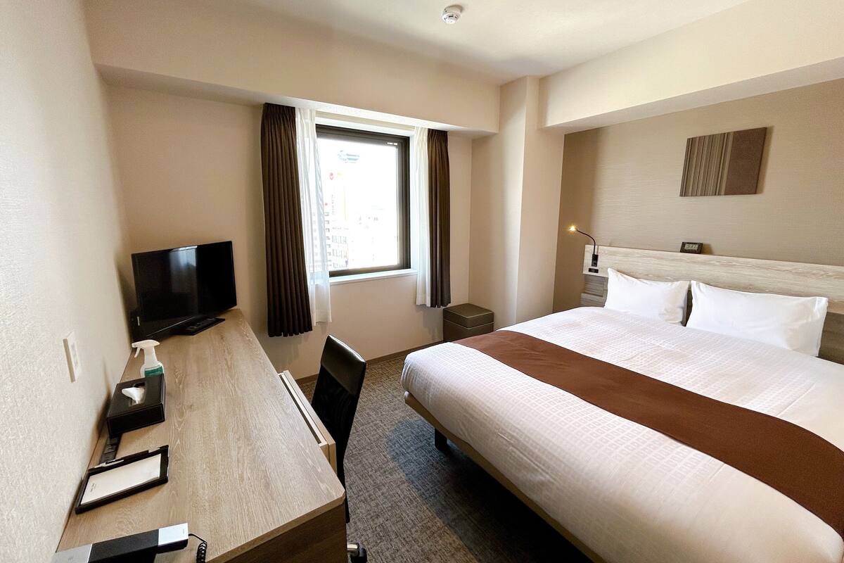 Una habitación de hotel con cama, escritorio con televisor, ventana con cortinas y mesita de noche.