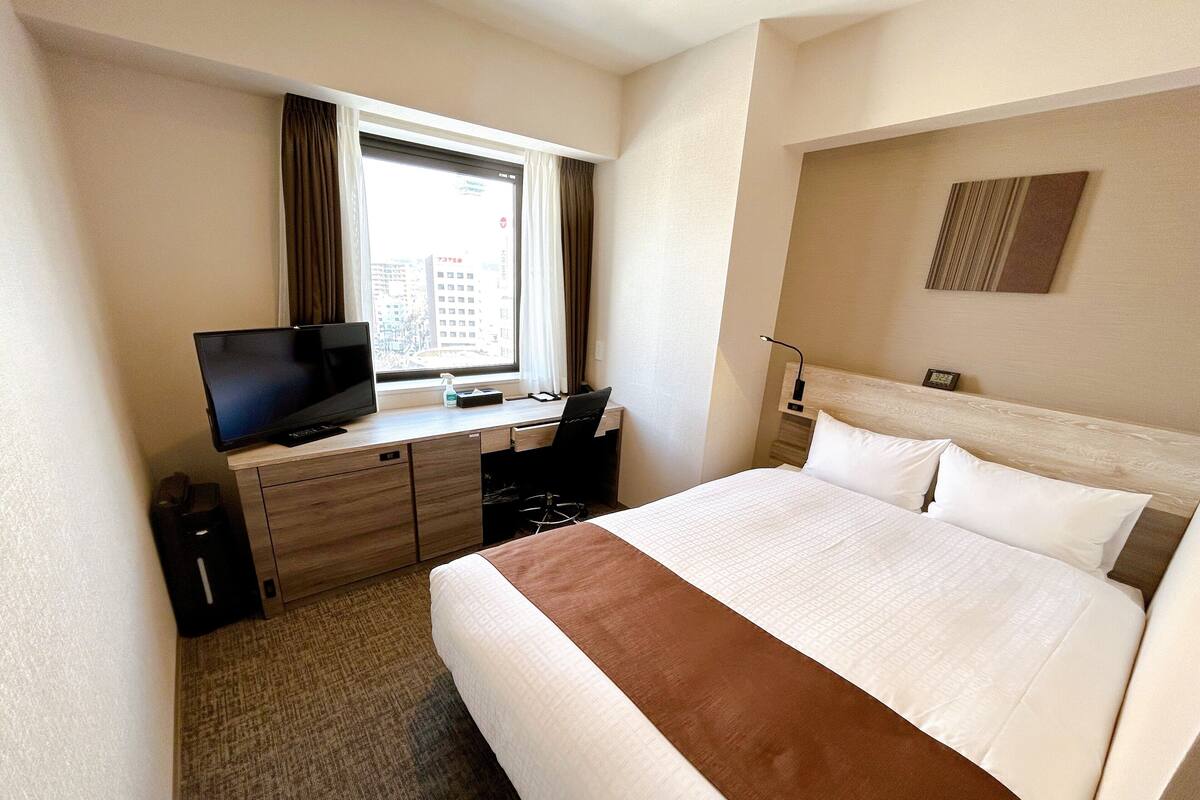Habitación de hotel con cama, escritorio con computadora, televisor y ventana con cortinas.