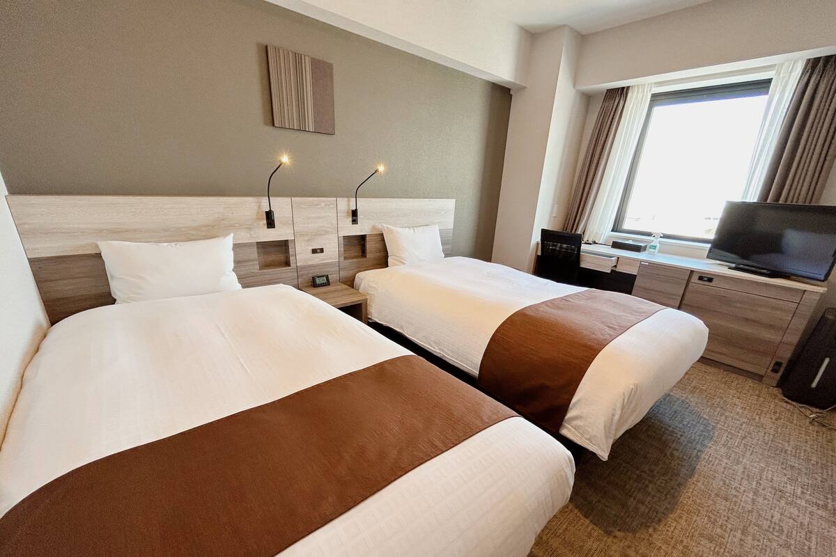 Habitación de hotel con dos camas, televisión, escritorio y ventana con cortinas.
