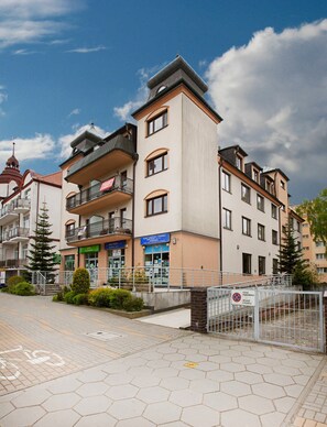 Un immeuble résidentiel à plusieurs étages, doté de balcons, d’une entrée bien visible et d’un espace de stationnement fermé par une grille.