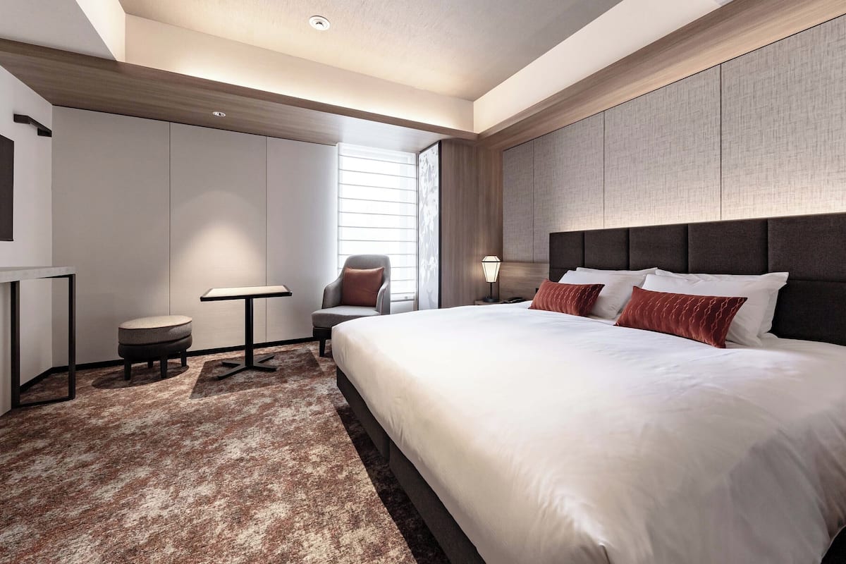 A modern hotel room with a large bed, a small table, a chair, and a nightstand with a lamp.