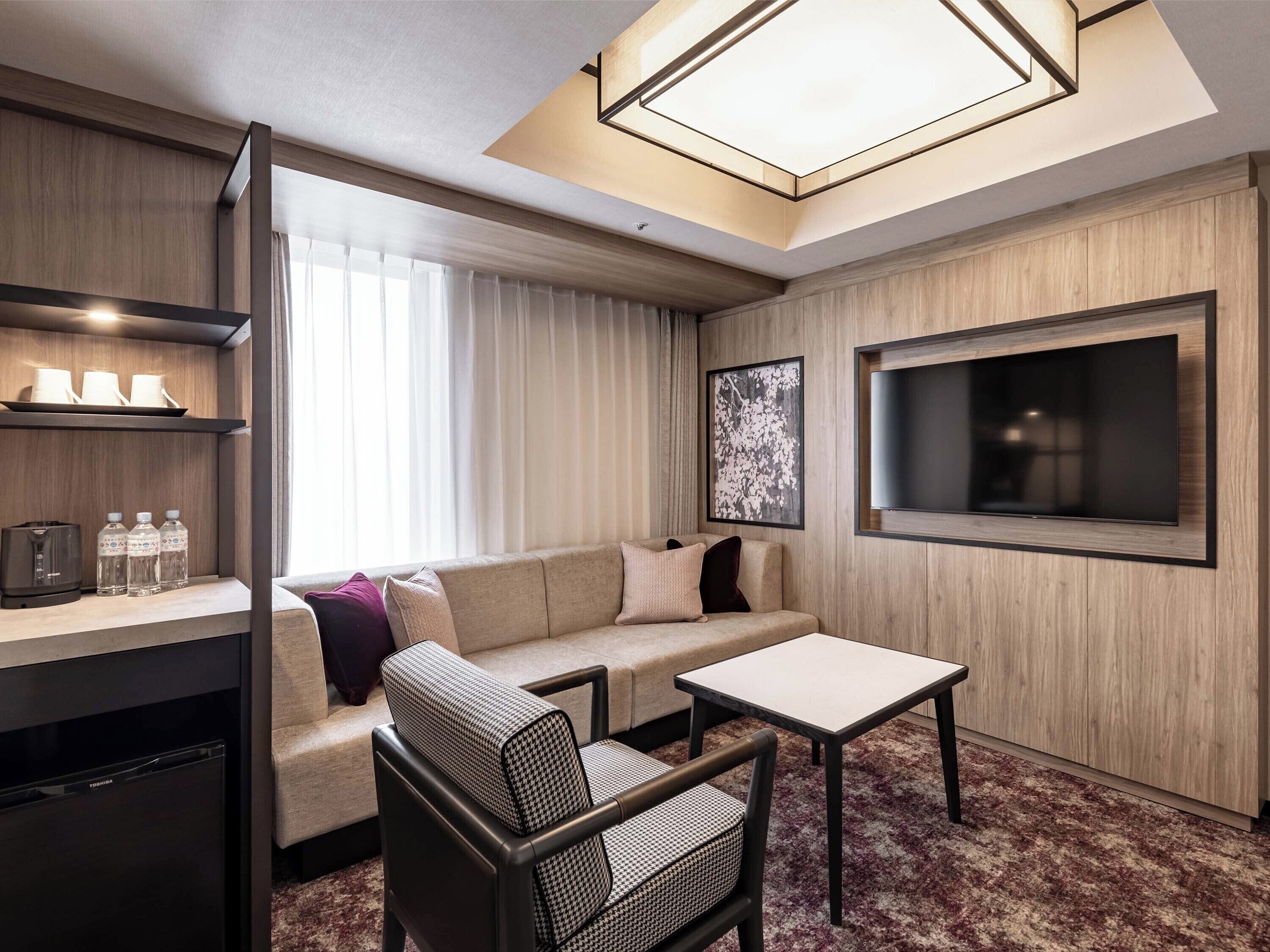 A modern hotel room with a sofa, armchair, small table, and a large TV.