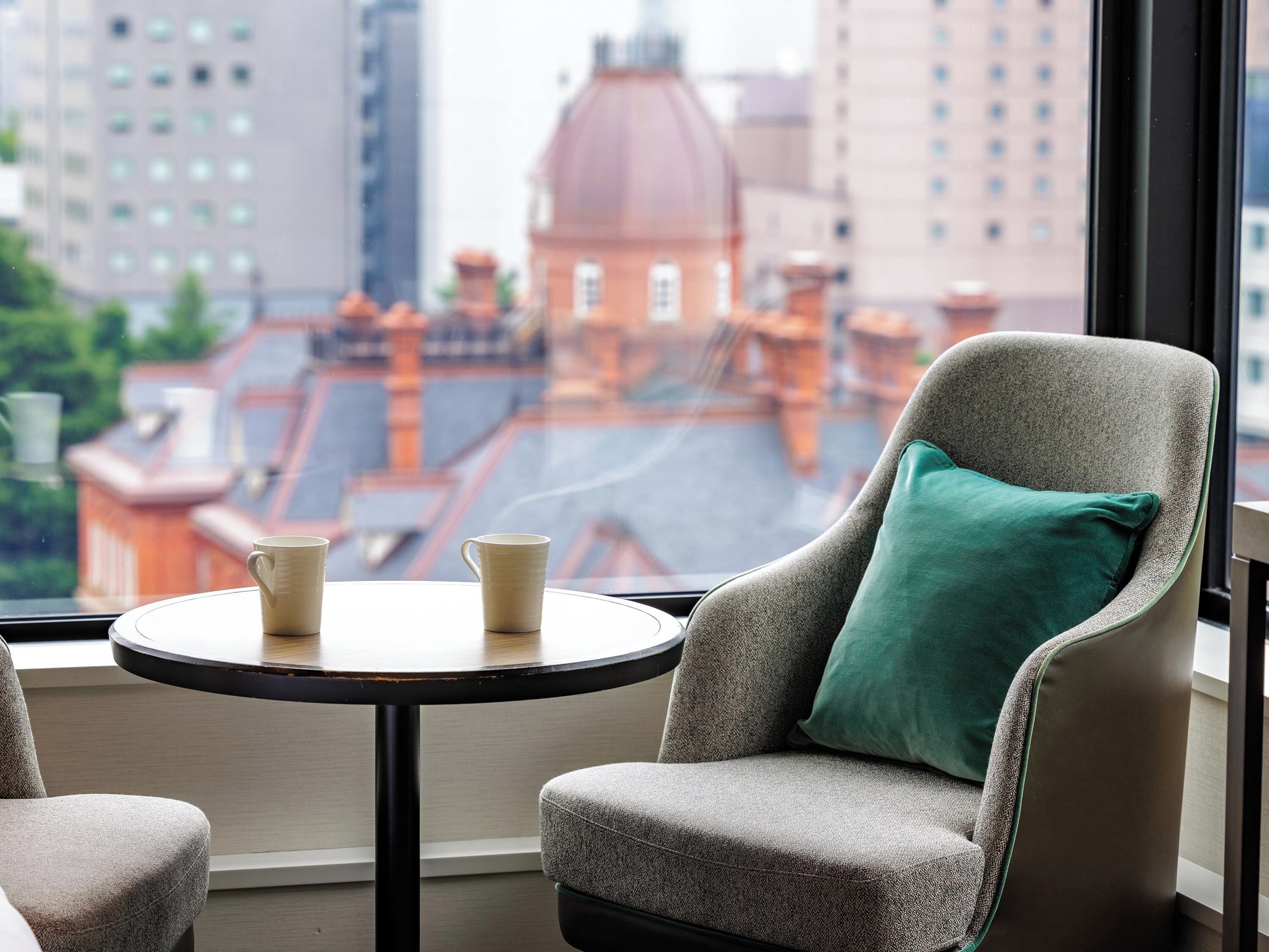 Une pièce avec vue sur le paysage urbain, une table ronde avec deux tasses et une chaise avec un coussin vert.