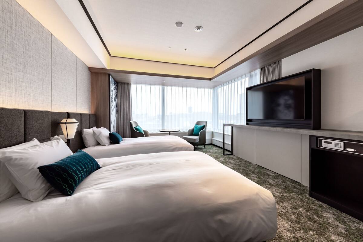 A modern hotel room with two beds, a TV, a desk, and a view of the city.