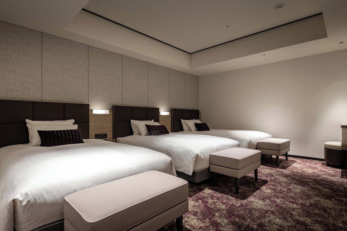 A modern hotel room with two beds, a large window, and a carpeted floor.