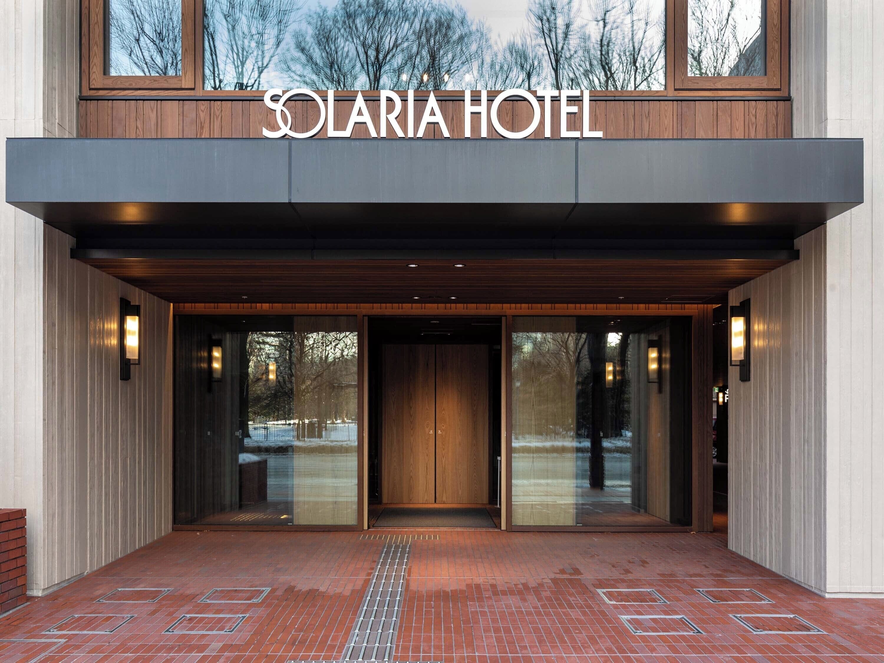 La entrada del Hotel Solaria con un diseño moderno, destacando un amplio acceso de madera, puertas de cristal y un camino de ladrillo que conduce a la entrada.
