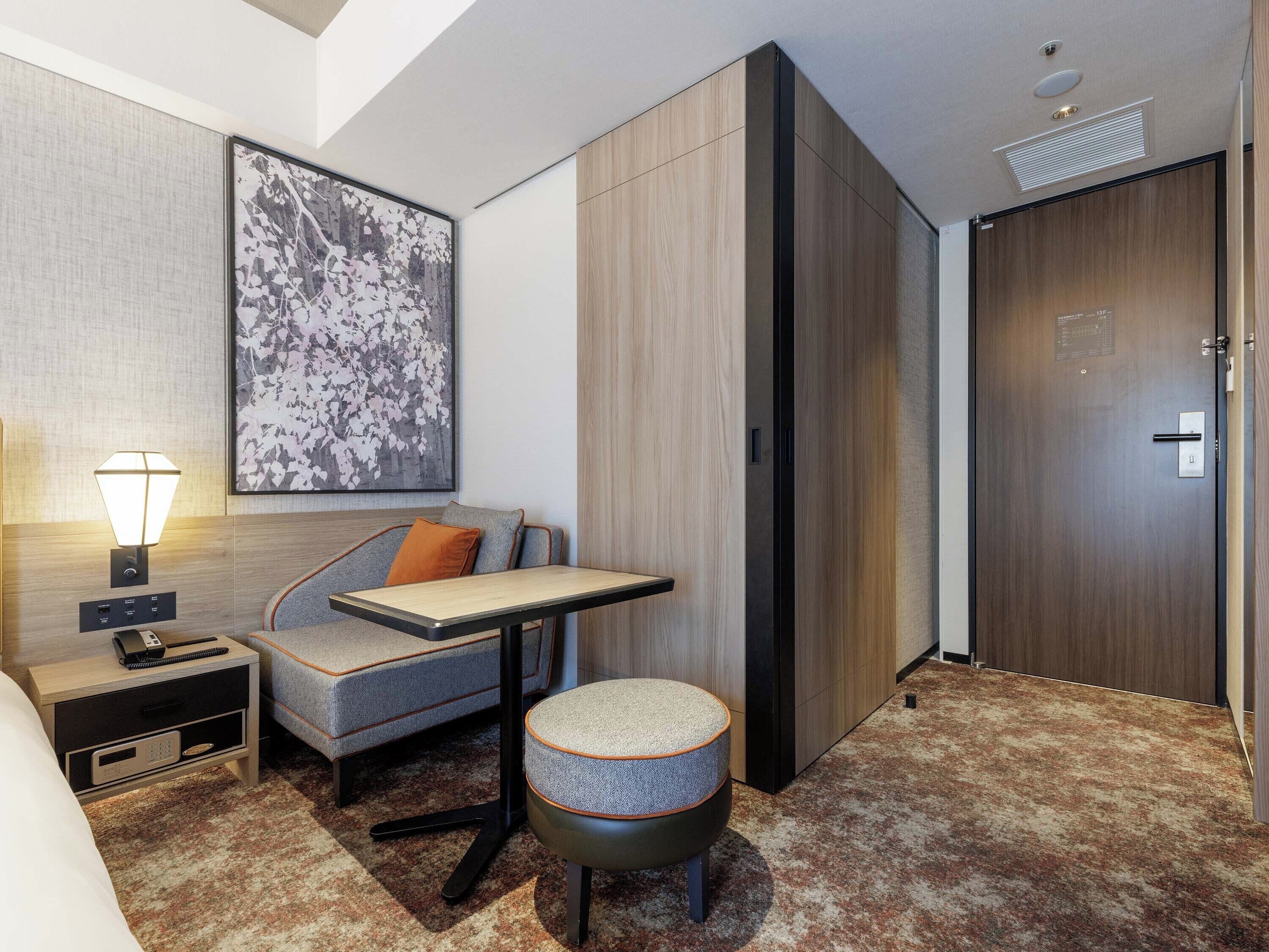 A hotel room with a bed, a desk, a chair, a nightstand, a lamp, a large abstract painting, and a wardrobe.
