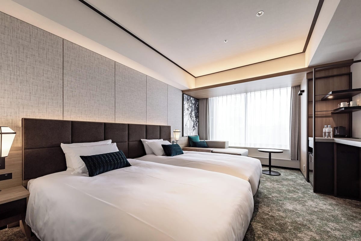 A modern hotel room with two large beds, a small seating area, a desk, and a large window with curtains.