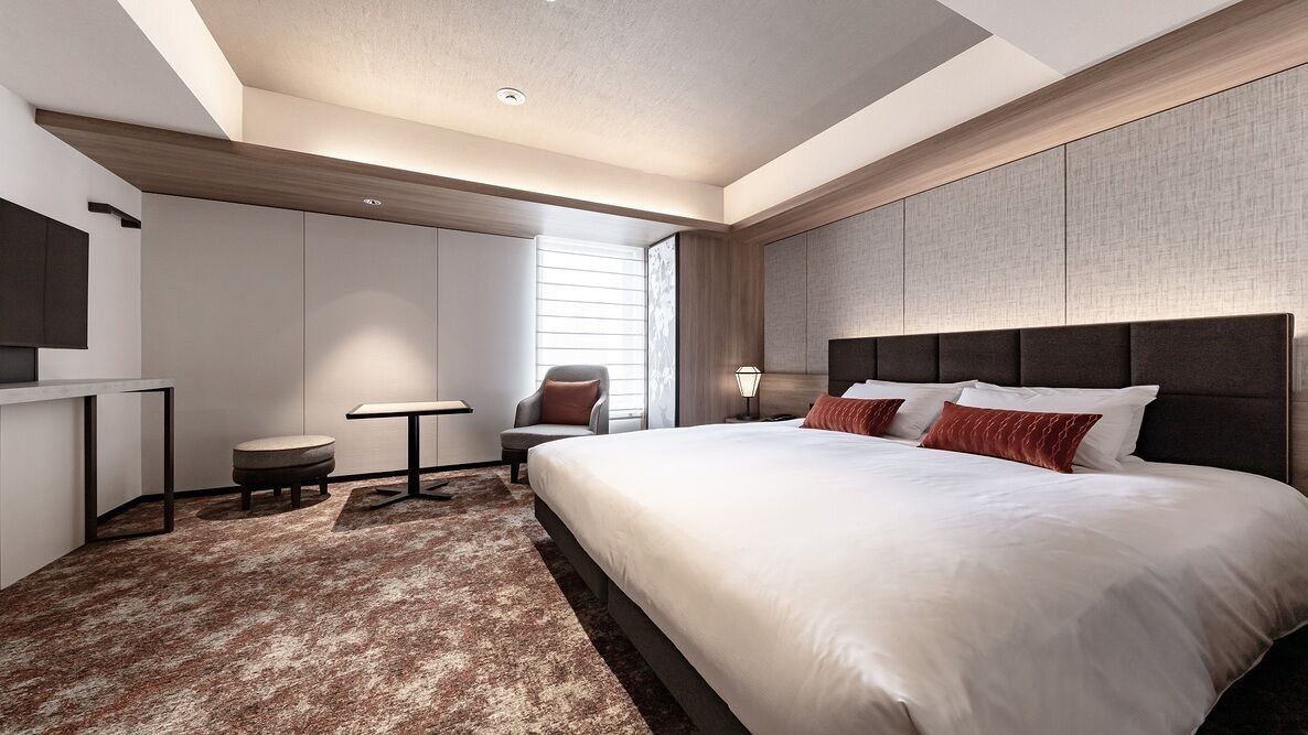 A modern hotel room with a large bed, a desk, a chair, and a small table.