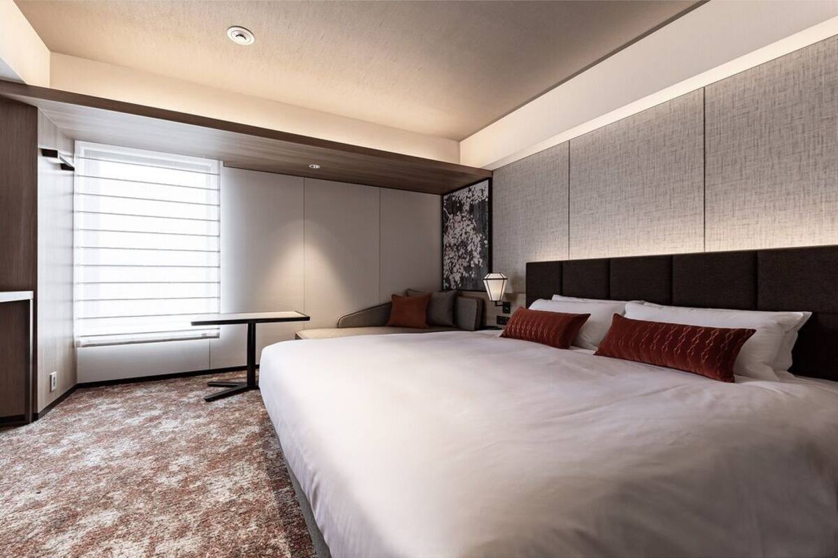 A modern hotel room with a large bed, a desk, and a window with blinds.