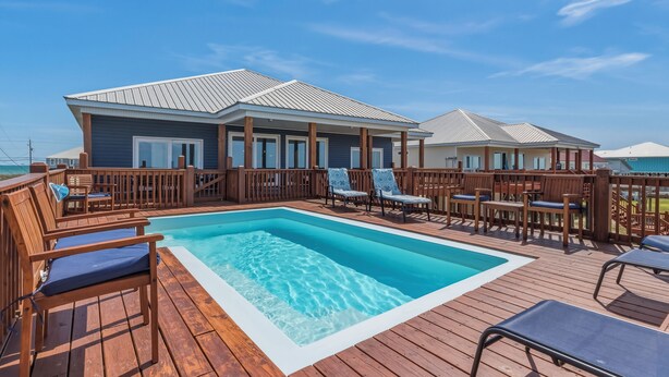 Un espace piscine avec une terrasse en bois et des chaises longues, une structure couverte dotée de fenêtres et un ciel d’un bleu éclatant.