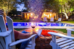 Una persona relajándose junto a una piscina, con una copa de vino, rodeada de mobiliario de exterior y un patio bien iluminado.