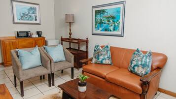 A living room with a brown leather sofa, two armchairs, a wooden coffee table with a plant, a wooden side table, a lamp, and a framed painting on the wall.