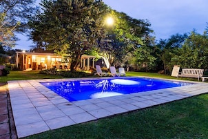 Una piscina con sillas de descanso, un área de estar techada y una casa iluminada al fondo.