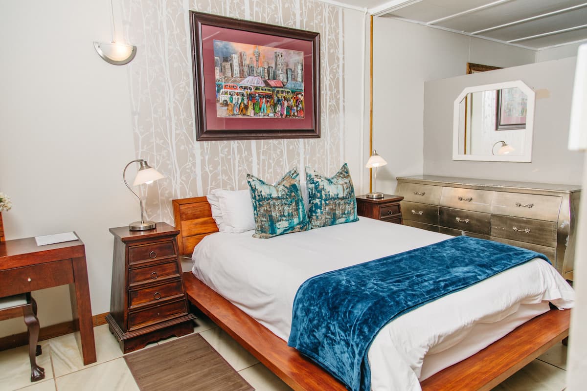 A bedroom with a wooden bed, white bedding, a blue throw blanket, a framed cityscape picture, and a nightstand with a lamp.