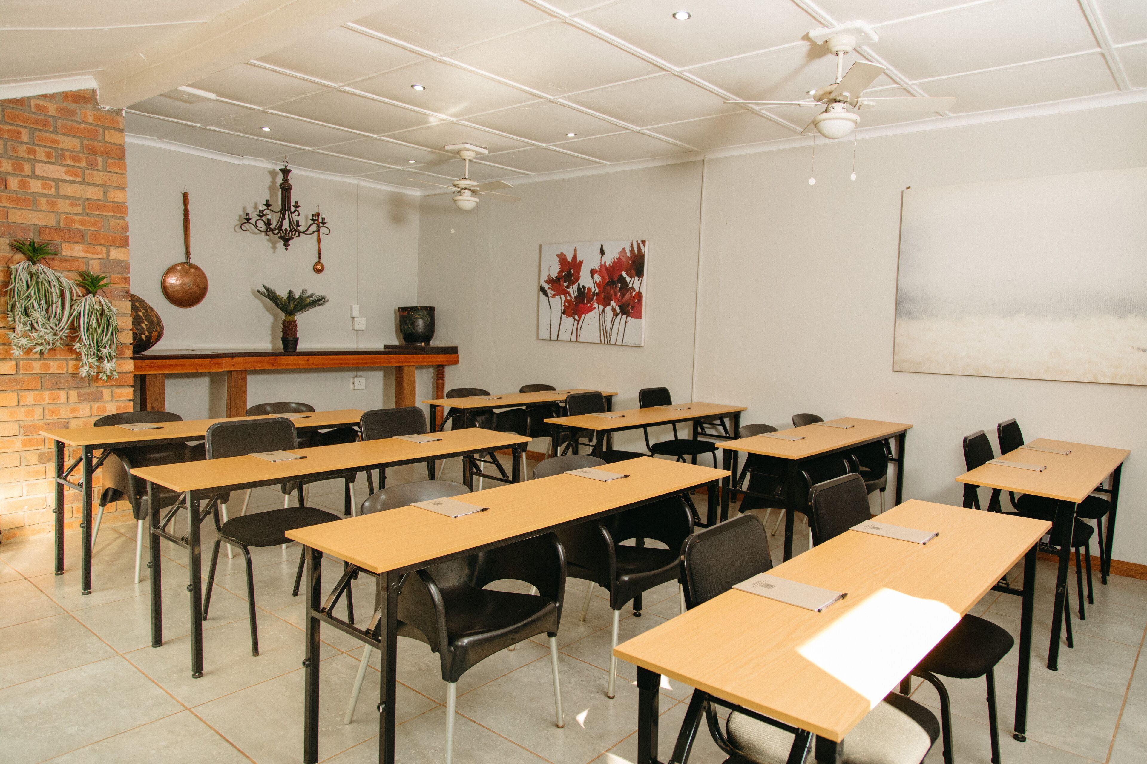 A classroom with wooden desks and black chairs, a brick wall, a chandelier, and a painting on the wall.