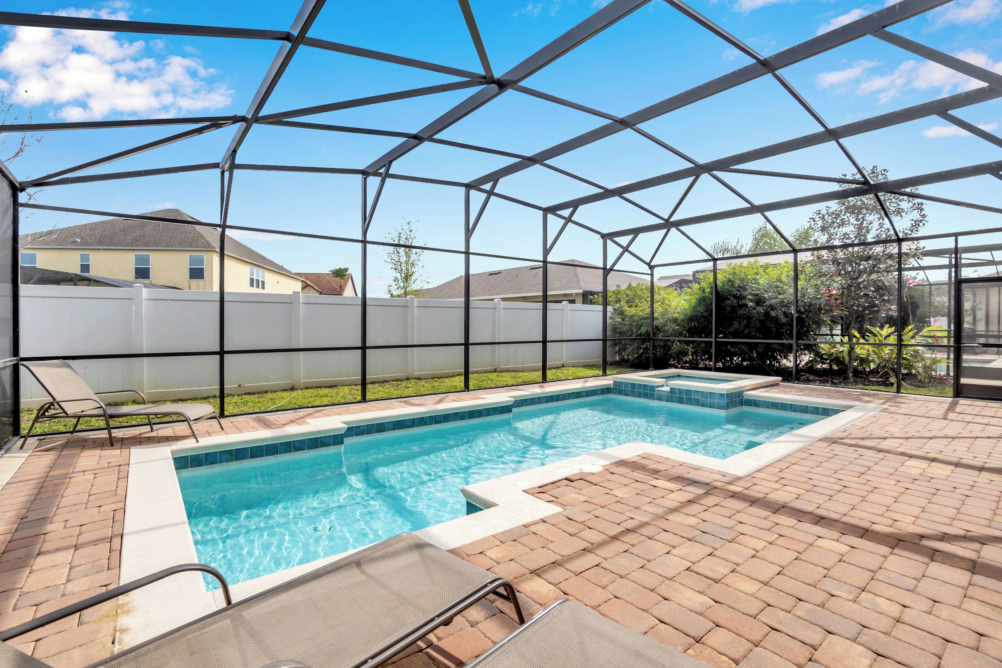 A covered outdoor swimming pool with a clear blue water feature, surrounded by a brick patio and a white fence.