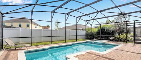 A covered outdoor swimming pool with a clear blue water feature, surrounded by a brick patio and a white fence.