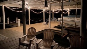 An outdoor deck with wooden chairs and tables, string lights, and a boat in the background. - Just One More Day: 2 Kayaks, Wifi, Horseshoes, Paddleboard, Panoramic View (Huddleston)
