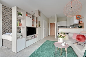 Sala de estar moderna com televisão de tela plana, prateleiras embutidas, tapete verde, mesa rosa e área de cozinha ao fundo.