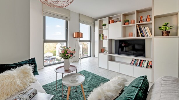 A modern living room with a large TV, white shelving, and a green rug. - Flats For Rent - Garden Gates (Gdansk)