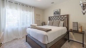 A bedroom with a large bed, a bedside table, a nightstand, a window with sheer curtains, and a wall-mounted light fixture. - The Ranch - Private Pool / Games / Outdoor Fire pit- 3BD / Huge Office (Phoenix)