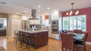 A modern kitchen with a central island, bar stools, a dining table with chairs, and a large window with blinds. - The Ranch - Private Pool / Games / Outdoor Fire pit- 3BD / Huge Office (Phoenix)