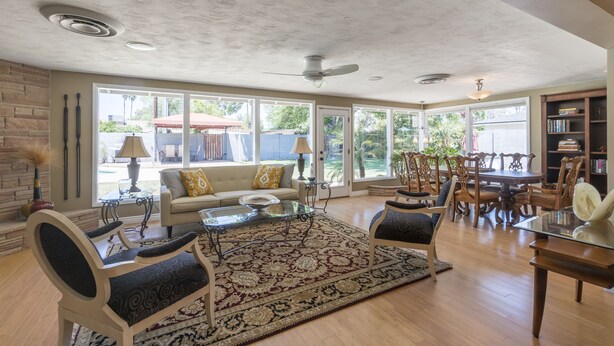 A spacious living room with a large window, a ceiling fan, and a dining area with wooden chairs and a table. - The Ranch - Private Pool / Games / Outdoor Fire pit- 3BD / Huge Office (Phoenix)