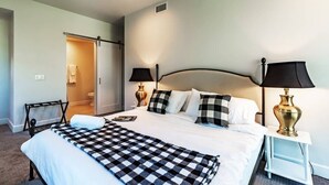 Habitación de hotel con cama, dos lámparas de noche, una manta de cuadros y una puerta que da al baño.