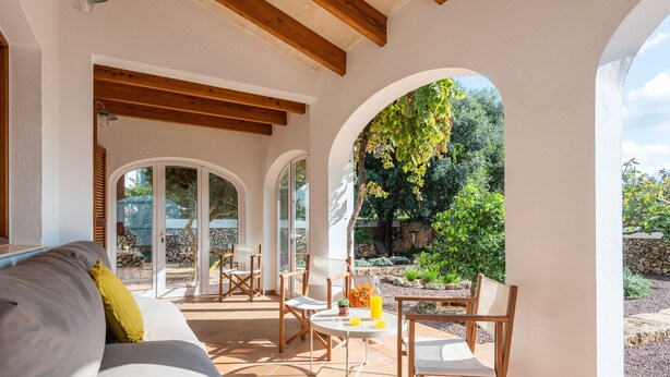 Un patio soleado con sofá, sillas y una mesa, que ofrece vistas a un jardín y un muro de piedra.