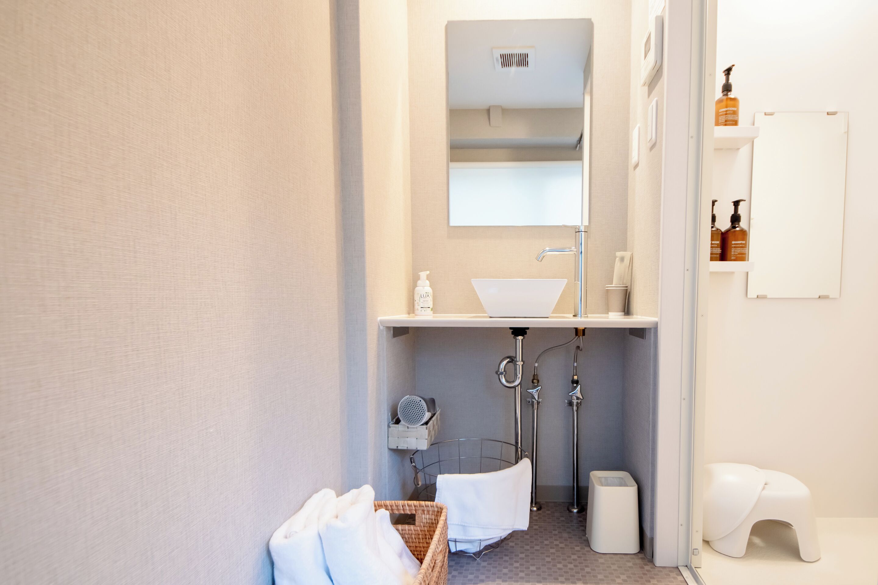 A modern bathroom with a white sink, mirror, and towel rack. There is a small trash bin and a white stool.