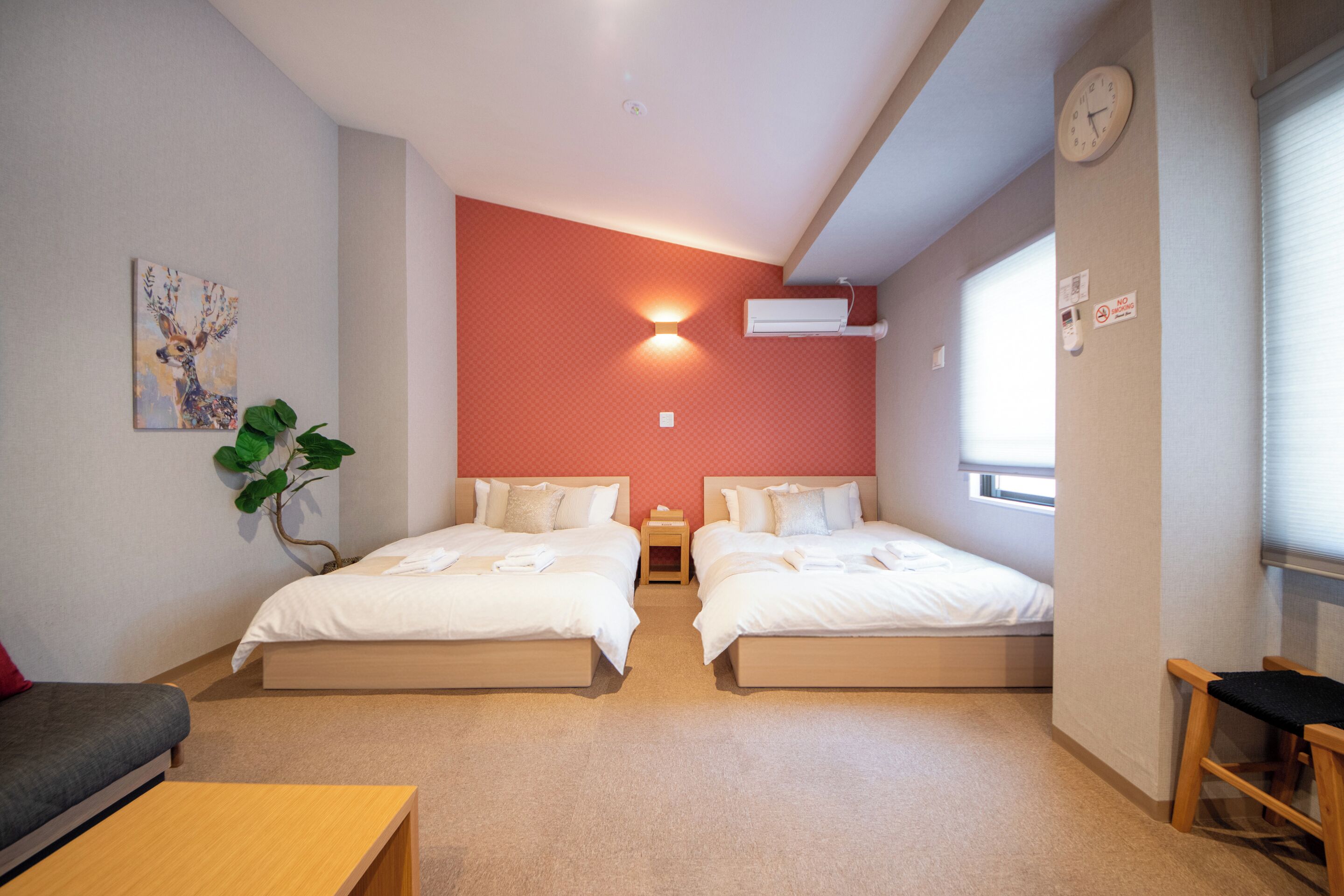 A room with two beds, a sofa, a small table, a wall clock, a wall-mounted air conditioner, and a window with blinds.