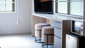 Une chambre moderne équipée d’un téléviseur à écran plat posé sur un bureau en bois, de deux tabourets rembourrés et d’un petit réfrigérateur.