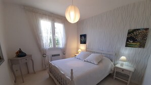 A bedroom with a bed, two bedside tables, a window with curtains, a wall-mounted painting, and a hanging light fixture. - Cozy House with Garden near Thermes du Connétable - 2 BR, Fully Equipped, WiFi, Sleeps 4 (La Roche-Posay)