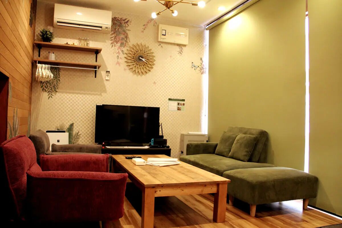 A living room with a wooden table, a red armchair, a green sofa, a flat-screen TV, and decorative wall art.