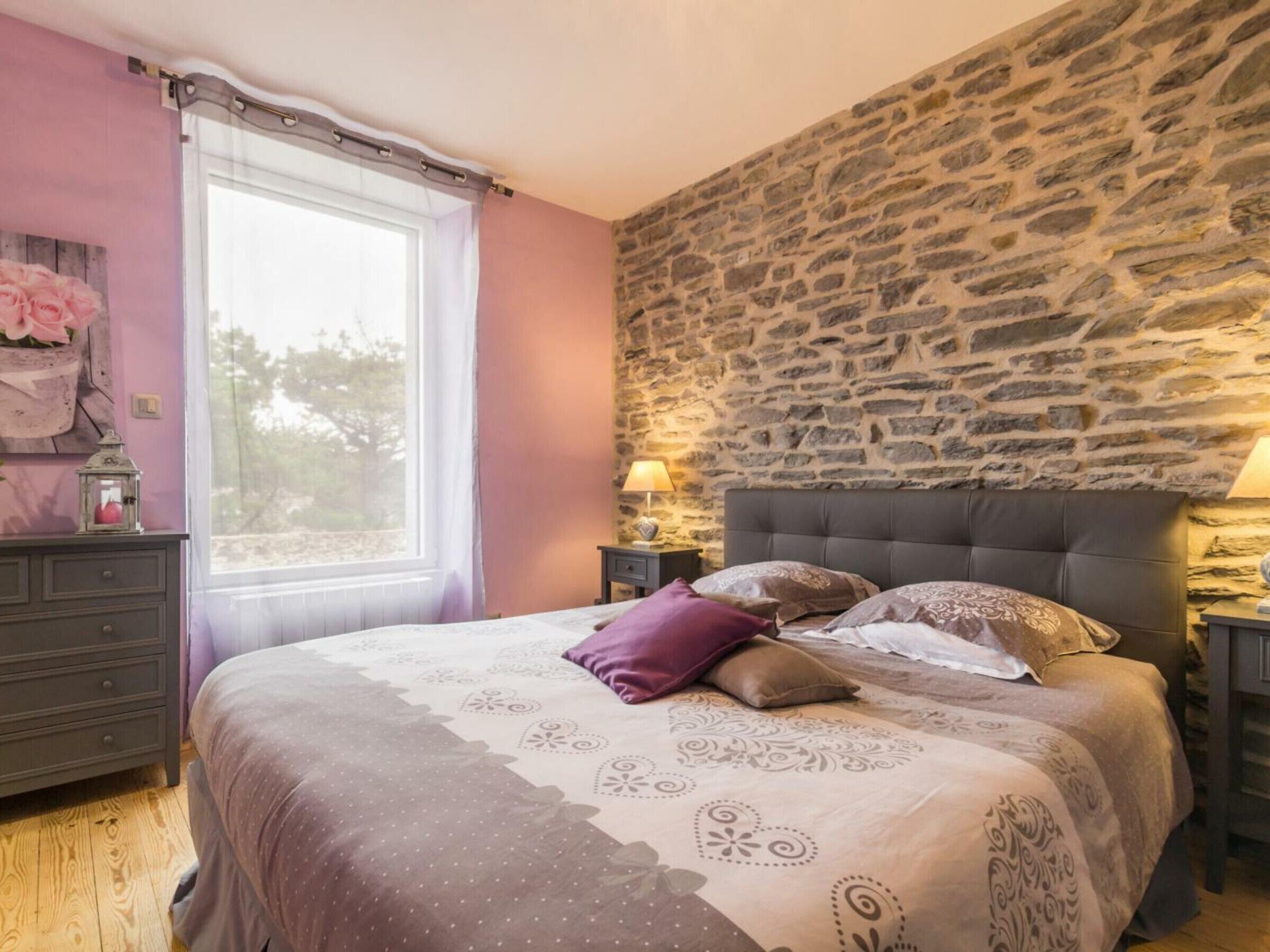 Un dormitorio con pared de piedra, cama con ropa de cama estampada, cómoda y ventana con cortinas.