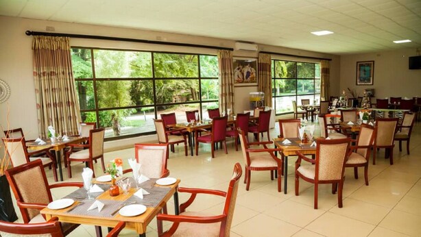 A spacious dining area with multiple tables and chairs, large windows offering a view of greenery, and a ceiling fan. - Fatmols lodge (Ndola)