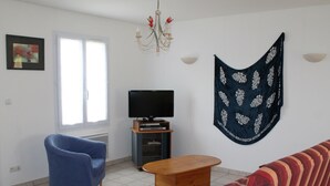 Un salon avec un fauteuil bleu, une table basse en bois, un canapé à rayures rouges, un téléviseur posé sur un meuble et une tapisserie décorative.