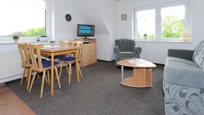A room with a dining table, chairs, a television, a sofa, and a window with a view of greenery. - Ferienwohnung 1 im Ferienhaus Trautes Heim - Ferienwohnung 1 (Esens)