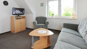 A living room with a television, a wooden cabinet, a grey armchair, a round table, and a grey sofa. - Ferienwohnung 1 im Ferienhaus Trautes Heim - Ferienwohnung 1 (Esens)